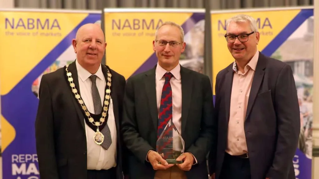 Guy Holden of Edenbridge Town Council holds a glass award between NABMA president Councillor David Hopkins, wearing a chain of office, and Mark Parry of sponsor ZAPP, in front of yellow and purple NABMA banners.